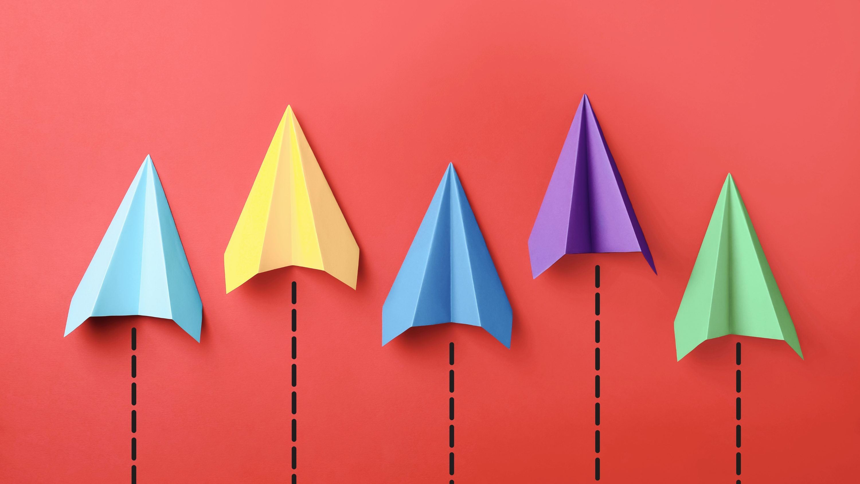 Quatre avions en papier de couleurs différentes (bleu, jaune, violet et vert) sur un fond rouge.